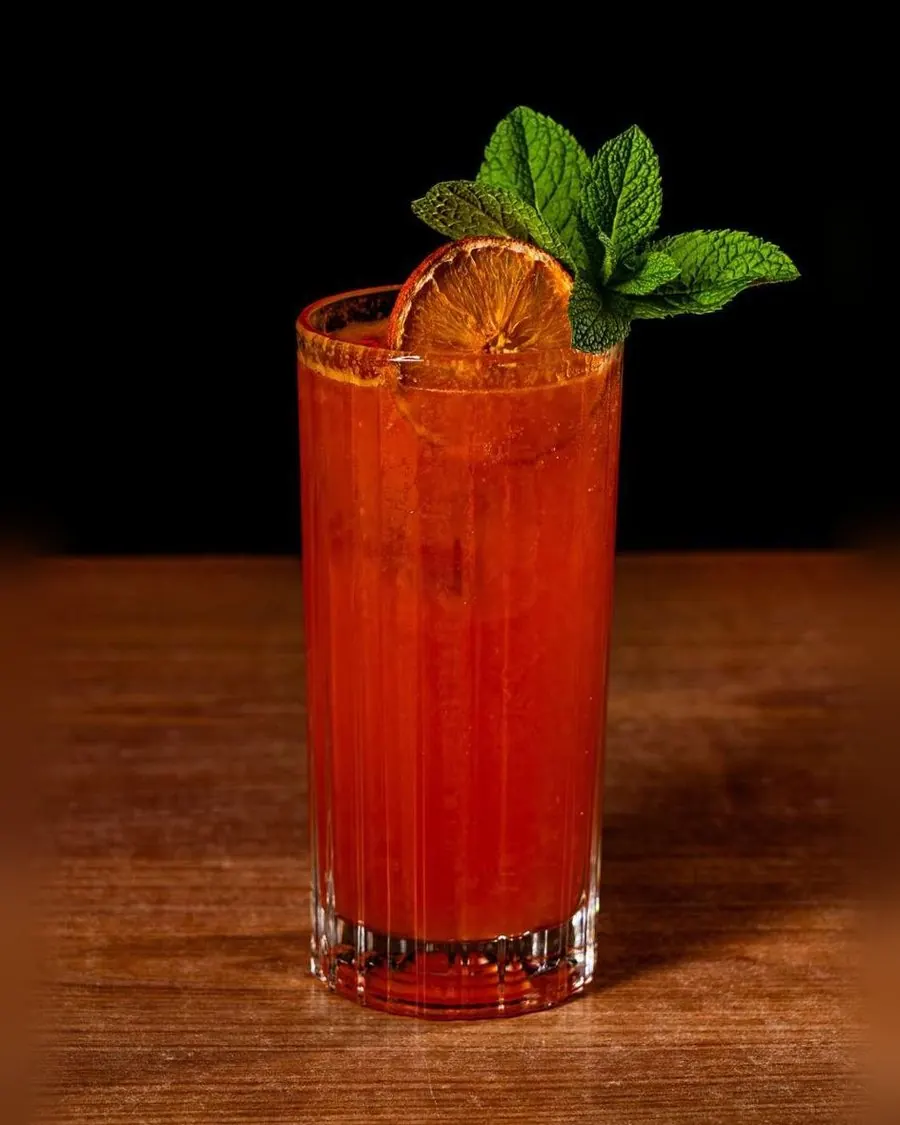 Cóctel Sunset rojo con naranja deshidratada y hierbabuena en vaso largo